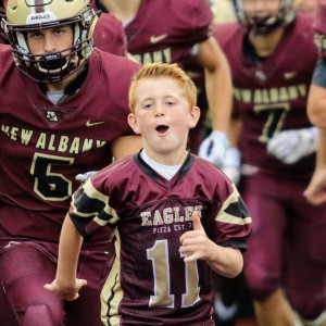 Young boy with a New Albany football jersey on running and cheering with the New Albany football team running behind him.