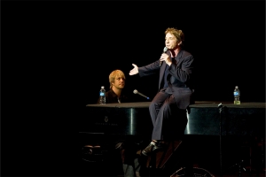 Entertainer Martin Short sitting on a piano, on stage with a microphone, performing at the McCoy's second year gala.