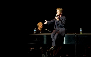 Entertainer Martin Short sitting on a piano, on stage with a microphone, performing at the McCoy's second year gala.