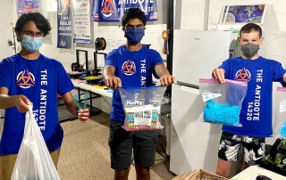 3 younger boys wearing face masks and holding up plastic bags of COVID-19 supplies.