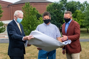 Three men with masks on standing outside reviewing a large sheet of blueprints.