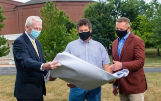 Three men with masks on standing outside reviewing a large sheet of blueprints.