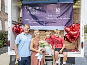 Angela Douglas holding flowers and posing behind an open cargo truck full of boxes with her husband and two sons.